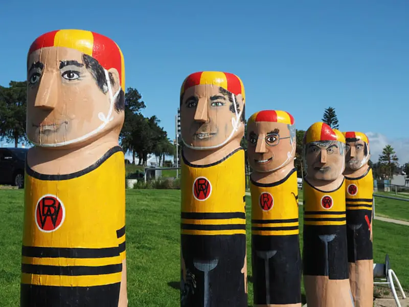 A row of colorful wooden bollards painted to resemble important people, each wearing a yellow and black striped jersey with a red and black emblem, standing on a grassy field under a bright blue sky.