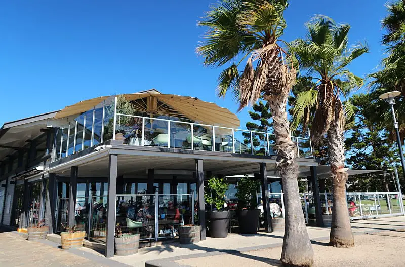 The Wharf Shed cafe, captured on a sunny day, boasts a modern architectural design with its large glass façade and angled roof. Positioned amidst tall palm trees, the establishment provides patrons both inside and on the balcony with panoramic views of the Geelong waterfront, enhancing the dining experience with its coastal charm.