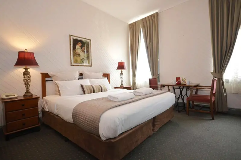 A cozy and well-appointed room at Central Springs Inn in Daylesford, featuring a plush queen-size bed with white linens and a tan comforter, flanked by wooden bedside tables and classic red-shaded lamps. A small desk and chair sit by the curtained window, under a vintage-style painting on the white wall.