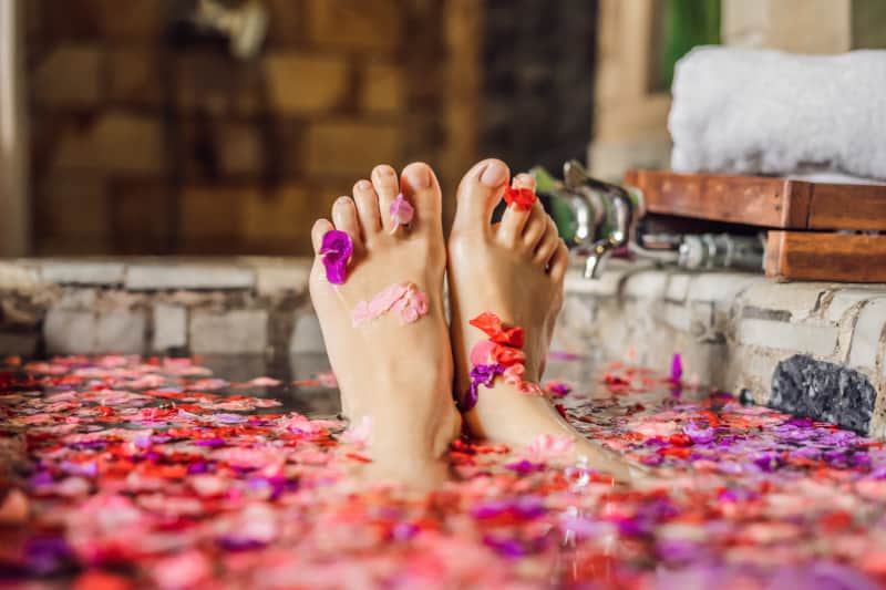 Feet adorned with colorful flower petals relax in a bath filled with a scattering of pink and purple petals, at the Hepburn Bathhouse and Spa, a haven for relaxation and rejuvenation.