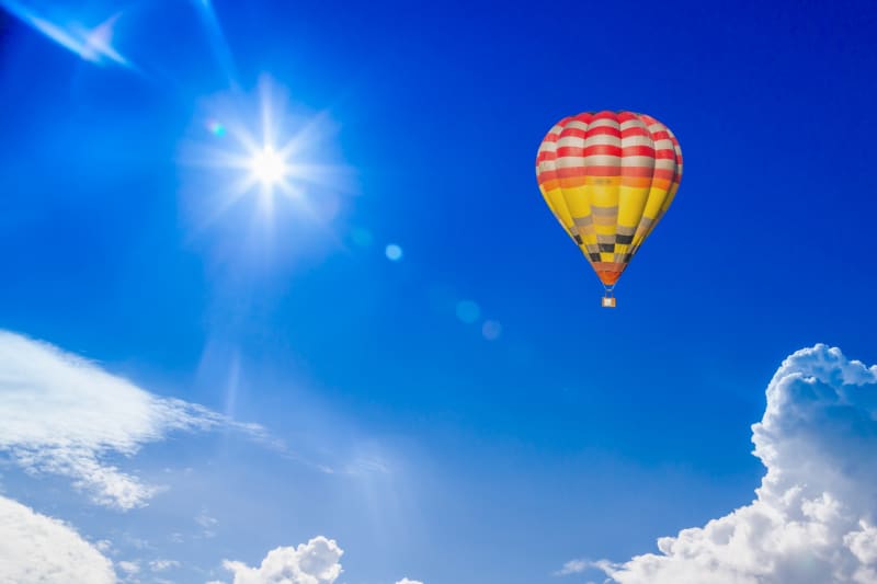 A vibrant yellow and red striped hot air balloon floats in a clear blue sky, with the sun casting rays and lens flare, suggesting a peaceful summer day high above the clouds.