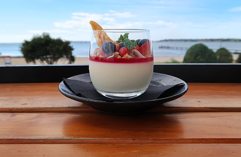 Breakfast smoothie at No 42 Geelong Waterfront Cafe, featuring a creamy base, red fruit coulis, fresh berries, and mint, served in a glass on a wooden table with a view of Rippleside Beach in the background.