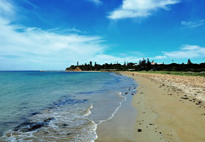 A panoramic view of Queenscliff Beach showcasing the serene blue waters gently lapping against the sandy shore, with the historic Black Lighthouse perched atop the distant cliffs under a clear sky.