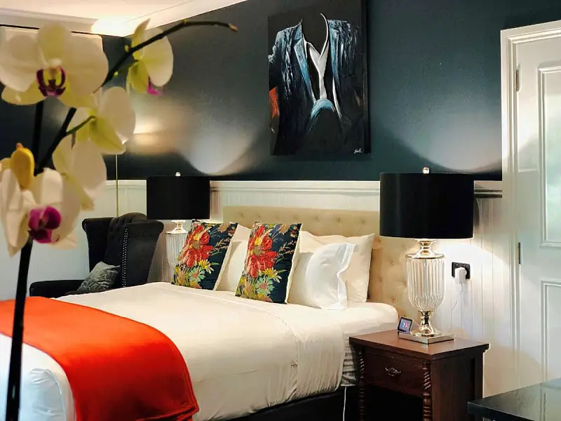 A tastefully decorated room at The Dudley Boutique Hotel in Daylesford, featuring a cozy queen bed with crisp white linens, accented by vibrant floral pillows, set against a dark teal wall with a sophisticated painting of a suit. A classic black lamp stands on the bedside table, while an orchid in the foreground completes the scene of luxury and comfort.