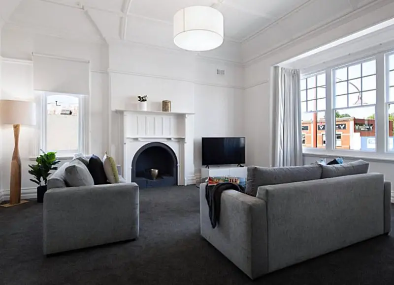 Spacious and elegant living room at 1913 On Vincent Daylesford accommodation, featuring plush grey sofas, a classic fireplace, and large windows with a view of the urban landscape outside.