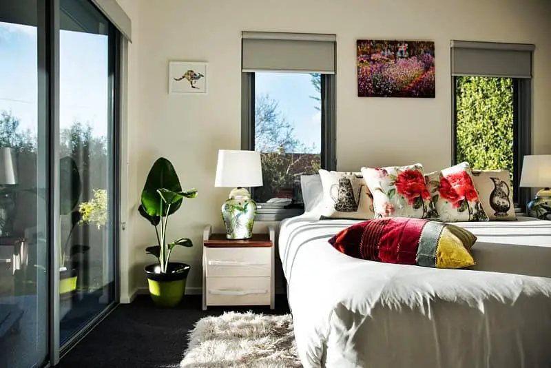 A modern and sunlit bedroom at Azura in Daylesford, featuring a comfortable bed adorned with colorful floral and bird-themed pillows, a fiddle leaf fig in a vibrant green pot, and artwork reflecting the natural beauty outside the sliding glass door leading to a lush garden.