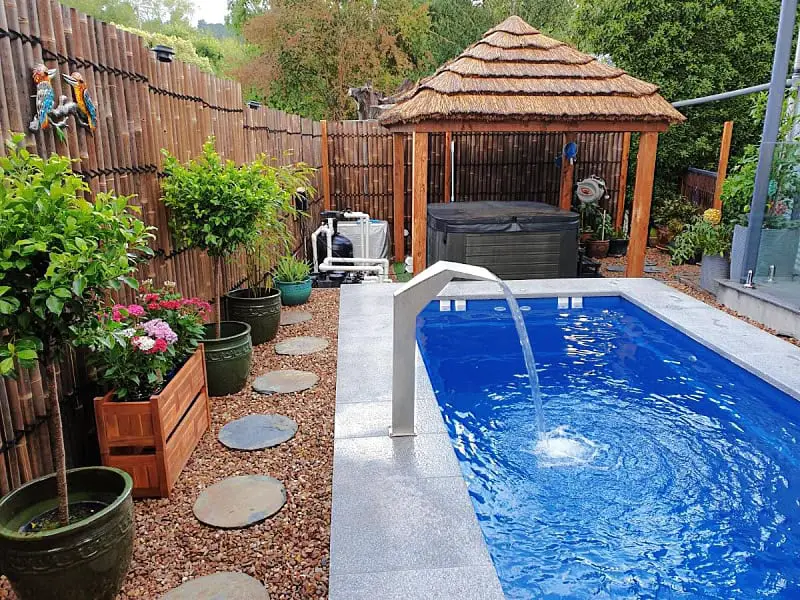 A tranquil outdoor pool area at Azura Daylesford luxury accommodation, with clear blue waters and a sleek water feature, complemented by a thatched-roof hut, hot tub, and lush potted plants along a pebbled walkway, creating a serene backyard oasis.