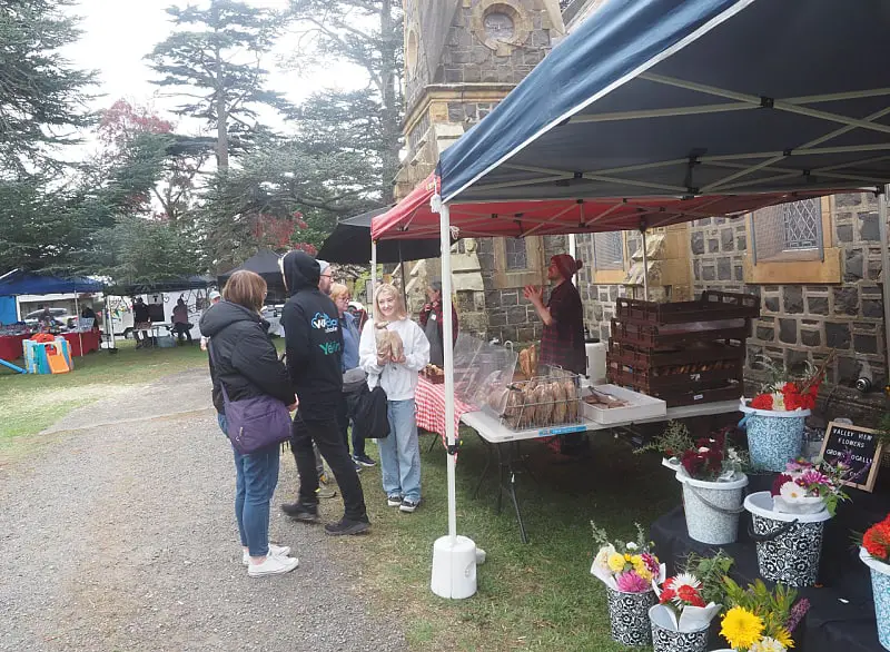 Visitors engaging with a vendor at Ballan Farmers Market, under a red and blue canopy beside a historic stone building. Fresh bread is displayed on a table, with vibrant potted flowers for sale, creating a lively local shopping atmosphere.