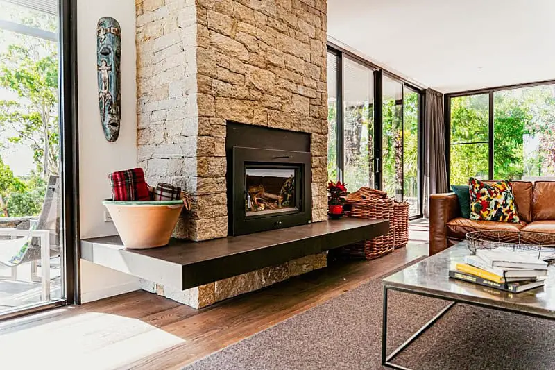 Cozy living room inside Bohemia Daylesford with a stone fireplace, leather sofas, and colorful cushions, surrounded by large windows offering a view of the green outdoors.