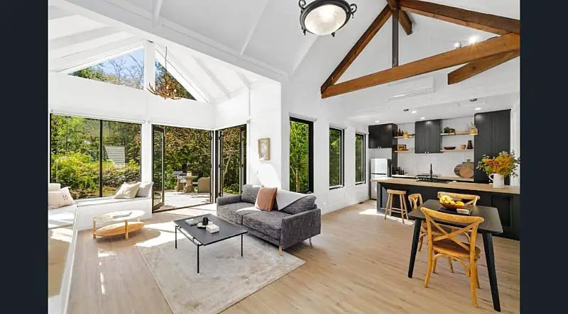 Bright and airy living space inside Daylesford Lake Garden Villas, featuring high vaulted ceilings with wooden beams, white walls, and large windows overlooking greenery, seamlessly integrating the living area with a modern kitchen and dining space.