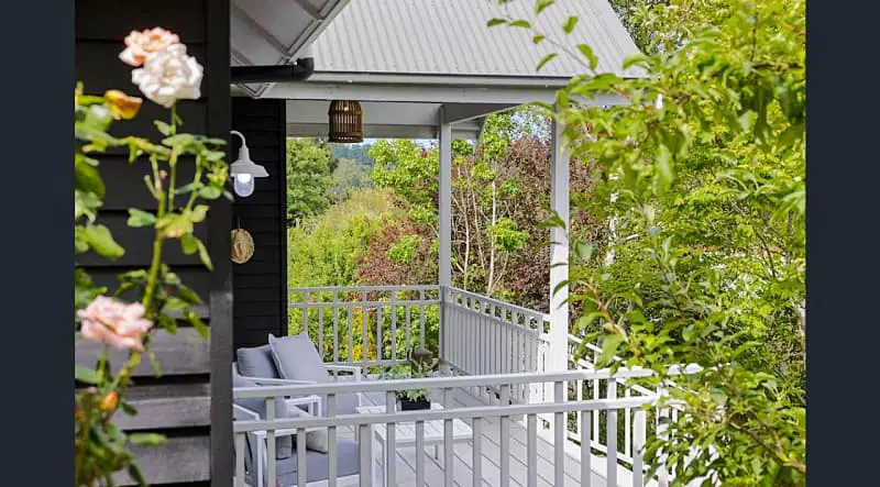 The cozy corner of a Daylesford Lake Garden Villa, featuring a well-appointed porch with white railings, comfortable seating overlooking lush greenery, and a hanging wicker lamp, framed by blooming roses and vibrant foliage.