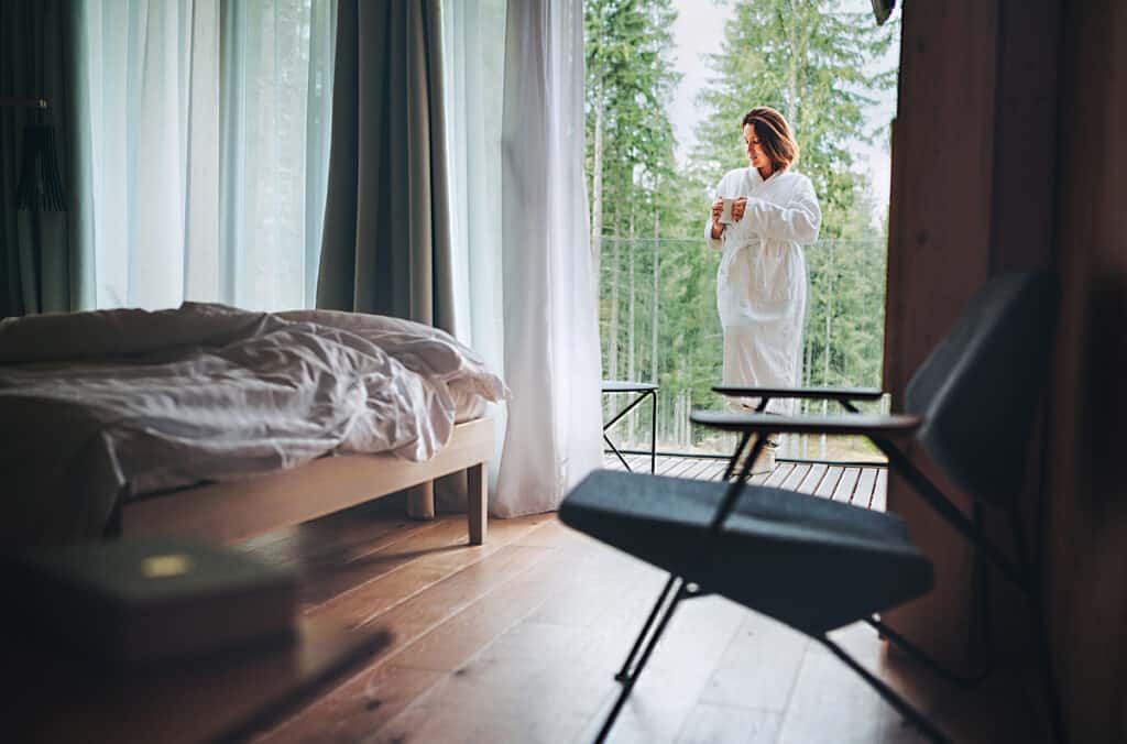 A serene morning scene at a Daylesford luxury accommodation, with a woman in a white robe standing on a balcony immersed in the tranquility of a forest, holding a warm beverage, next to a minimalist bedroom with a tousled bed and sheer curtains diffusing soft light.