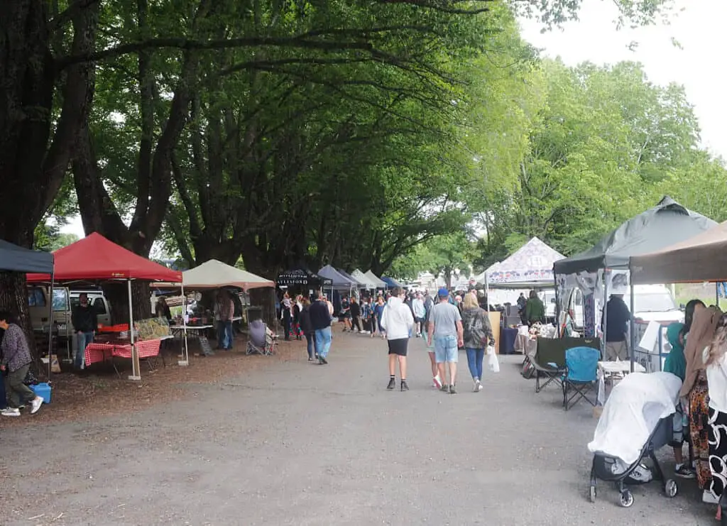 Visitors browsing various stalls at Daylesford Markets under a canopy of lush trees, with a mix of handmade goods and local produce on display.