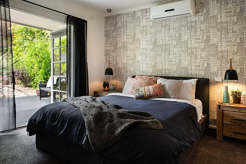 An inviting bedroom at East St 1 accommodation in Daylesford, with an open sliding door leading to a patio, showcasing a large bed with dark linen and a cozy gray throw, flanked by nightstands with modern black lamps, set against a wallpaper with a vintage building design.