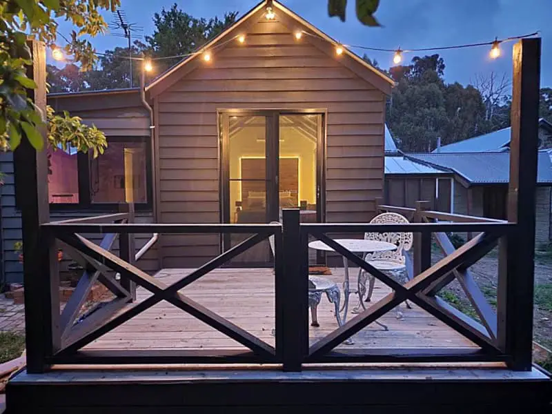 Twilight view of Frog Hollow Estate - The Cottage, showcasing its warm, glowing interior light that spills onto the wooden deck, where a vintage white metal chair invites relaxation under the string lights draped overhead.