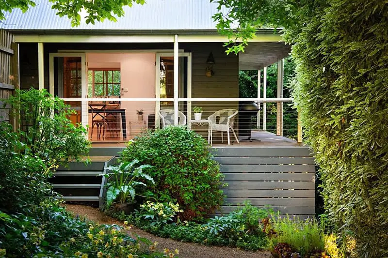 Tranquil garden setting of Haiku Daylesford with lush greenery surrounding a wooden deck and white chairs, leading to the cozy, open doorway of a country-style home.