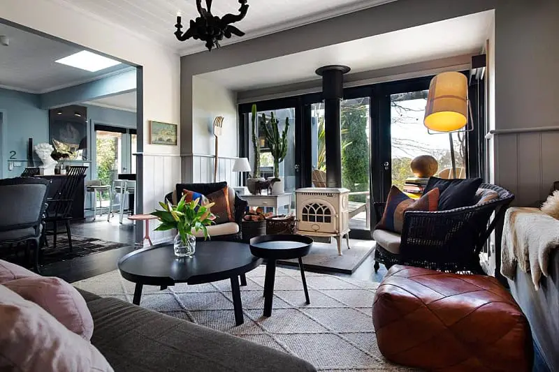 A cozy and stylish living room interior at Harts Lane Haus in Daylesford featuring a mix of modern and vintage elements. The space includes a black round coffee table, a cream-colored stove fireplace, large windows with garden view, and an assortment of comfortable seating options with decorative pillows and throws.