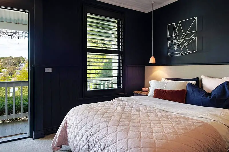 A cozy bedroom at Harts Lane Haus featuring dark walls and a large window with open blinds that provide a view of lush greenery. The room is elegantly decorated with a geometric wall art piece, a stylish pendant light, and a plush bed dressed in a mix of earth-toned linens and pillows.