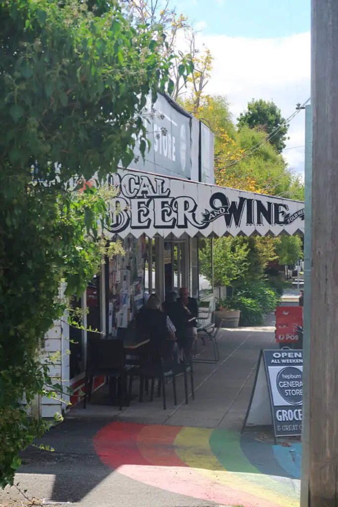 Outside the Hepburn General Store, patrons sit at tables under the shade of trees and a sign that reads 'LOCAL BEER AND WINE', with vibrant, multicolored sidewalk art leading the way.