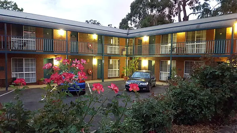 Twilight view of the Hepburn Springs Motor Inn with two levels of balconied rooms, vibrant pink flowers in the foreground, and a parked car facing the camera.