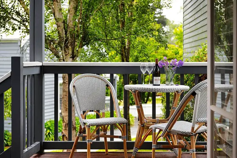 A cozy balcony setting at Lake Orchard Villas featuring two Parisian bistro chairs and a matching table, adorned with a vase of purple flowers, a bottle of wine, and two wine glasses, overlooking lush greenery.