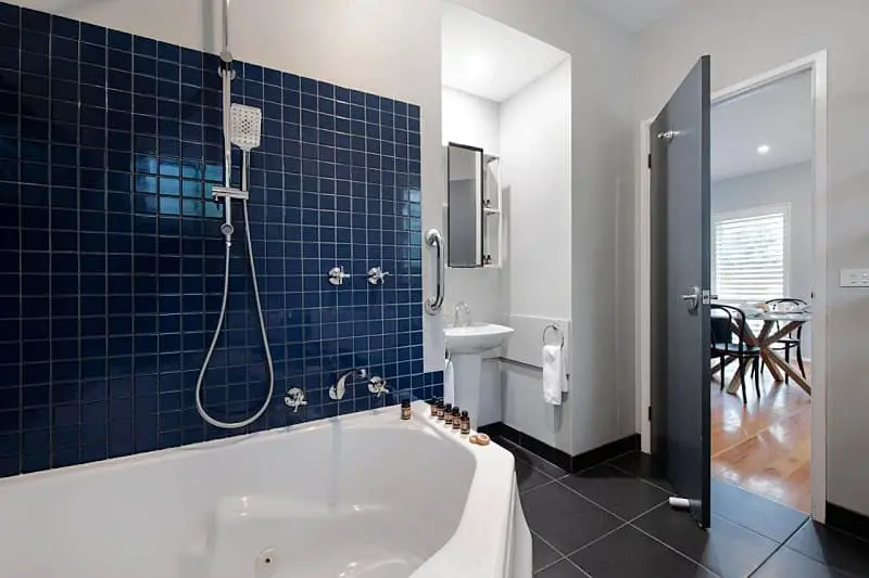 Elegant bathroom at Lake Orchard Villas with deep corner spa bath, dark blue tile surround, separate shower and classic white basin, viewed through an open door that offers a glimpse into the adjoining dining area.