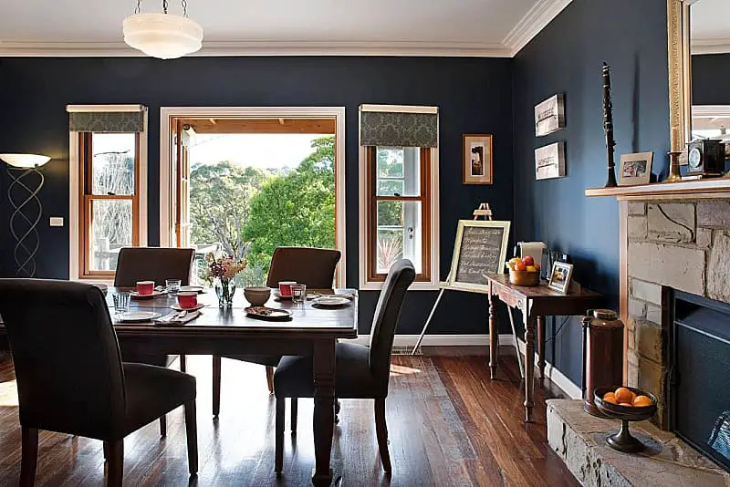 An elegant dining area at Old Chilli Daylesford bed and breakfast, with dark navy walls and hardwood floors. A classic wooden dining table set for a meal, flanked by comfortable chairs, sits by windows offering a view of the serene outdoors. Decor includes a fireplace with a rustic mantle, vintage decorations, and a chalkboard listing the day's specials.
