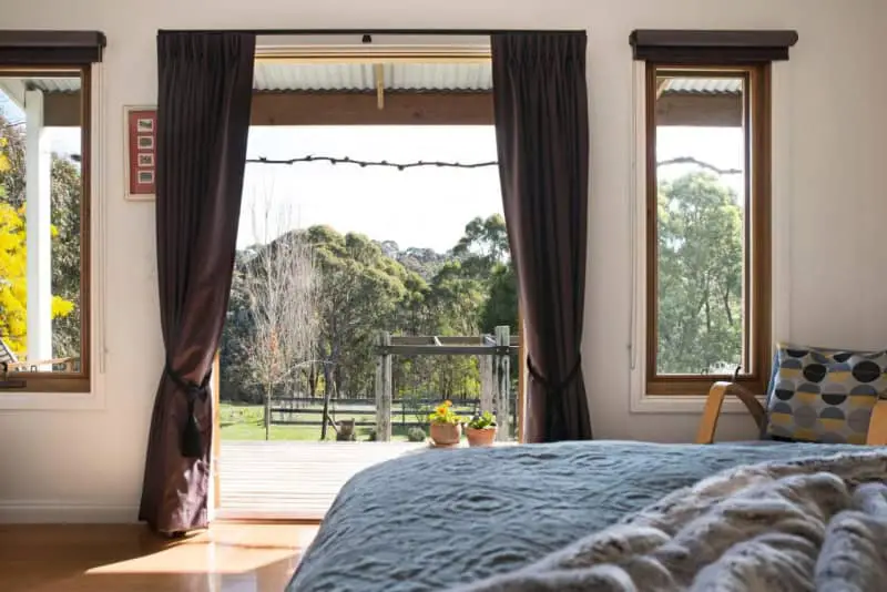 A cozy bedroom in Old Chilli Daylesford with a view of lush greenery through open French doors, leading to a sunny deck. Dark curtains frame the doorway, while a patterned chair and colorful cushions add a homely touch.