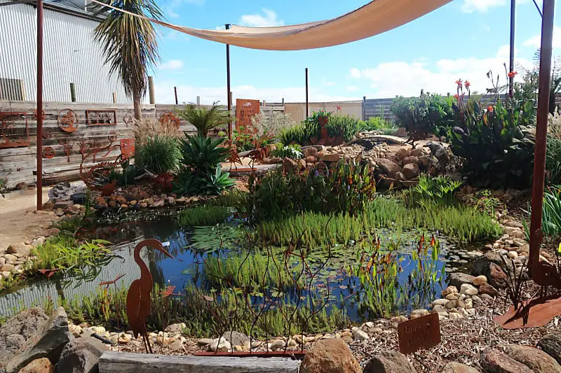 A tranquil water feature at the Overwrought Garden Art Gallery, adorned with rusted metal sculptures among vibrant greenery, under protective shade sails against a bright sky.
