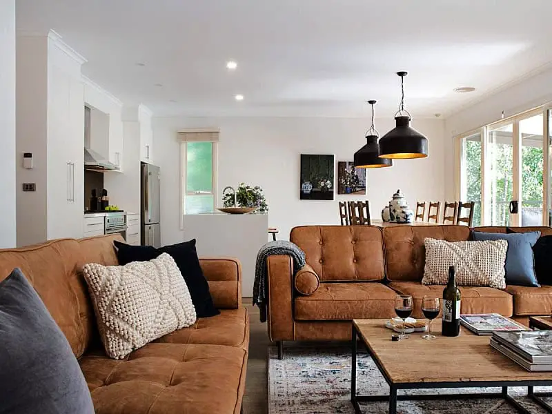 Cozy living area inside The Carlisle Daylesford holiday house, featuring plush caramel-colored sofas adorned with cushions, a wooden coffee table with wine glasses, and elegant pendant lights casting a warm glow.