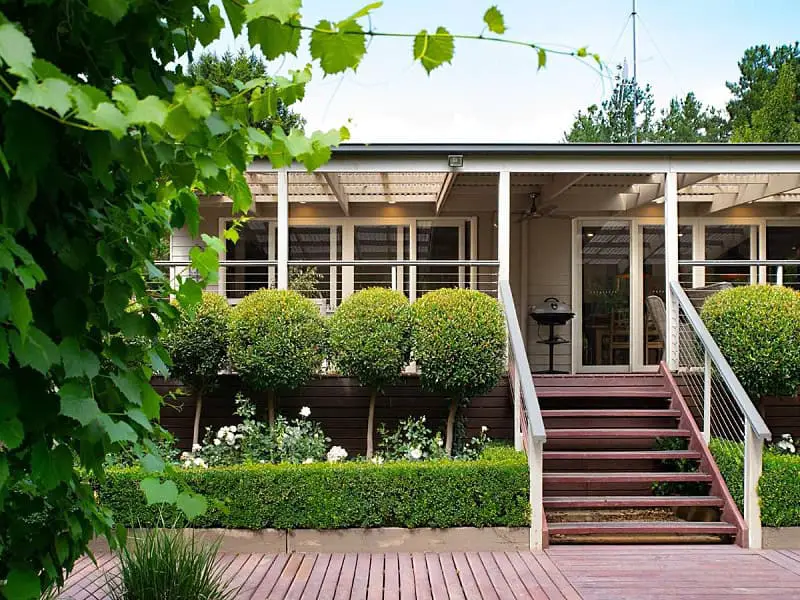 Charming view of The Carlisle in Daylesford, showcasing its serene garden with neatly trimmed round bushes, white blooming flowers, and a welcoming front porch with a staircase.
