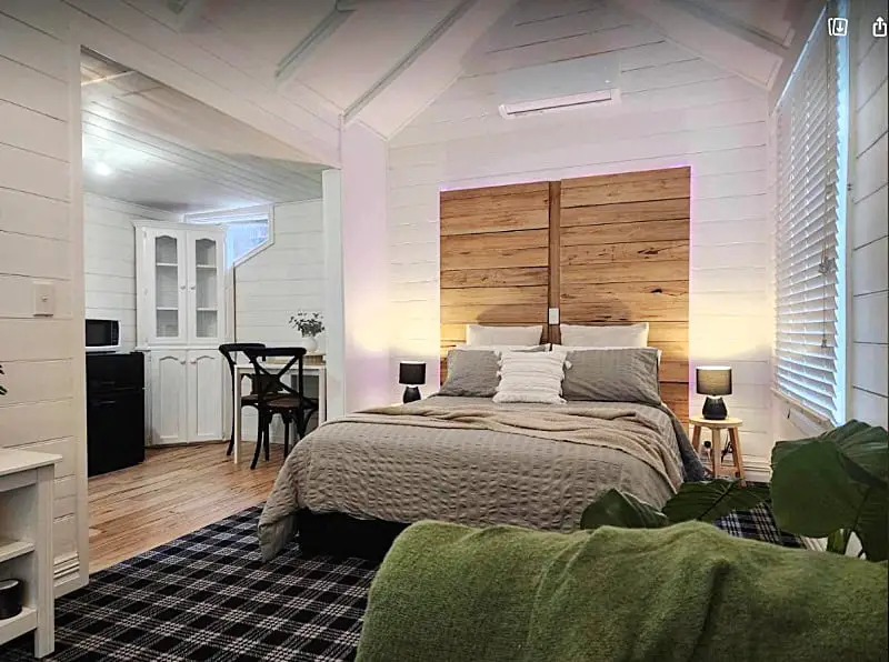 Cozy bedroom interior at Frog Hollow Estate - The Cottage, featuring a queen-size bed with plush linens, wooden headboard against white shiplap walls, a small kitchenette in the background, and ambient lighting creating a warm, inviting atmosphere.