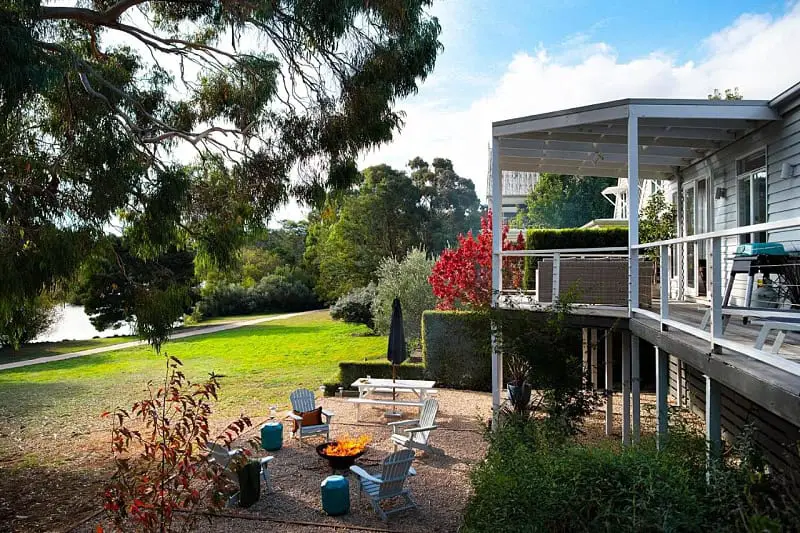 The Vue Lake Daylesford Villa showcases a serene outdoor seating area with a fire pit, set against a backdrop of lush greenery and a tranquil lake view, embodying a perfect blend of comfort and natural beauty.
