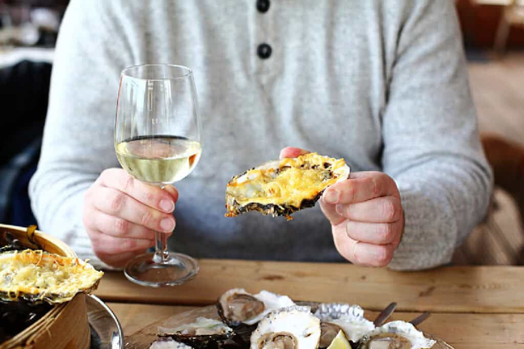 A patron at Fishermens Pier one of the best Geelong Waterfront Restaurants is depicted in a casual grey sweater, savoring the local cuisine with a grilled oyster in the left hand prominently shown to the camera. The right hand holds a glass of white wine, complementing the array of fresh oysters garnished with lemon wedges on the wooden table.