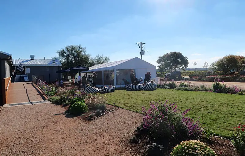 A sunny outdoor scene at Austin's Wines features a grassy area with people lounging on black and white striped bean bags next to a white tent. The setting includes a garden with blooming flowers, a wooden walkway leading to a building, and additional seating areas under umbrellas.