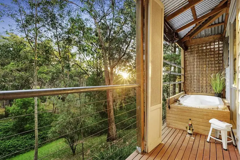Sunset view from the deck of Briars Cottage, showcasing a luxurious outdoor spa bath enclosed with bamboo privacy screens. The spa overlooks a lush, wooded area. A bottle of wine and a glass sit on a small white stool nearby.