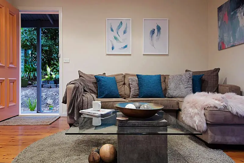 Interior of Briars Loft in Daylesford featuring a cozy living room with a plush sofa adorned with blue and gray cushions, a glass coffee table, and soft throw blankets. Artwork on the walls and a view of a garden through an open door add a serene atmosphere.