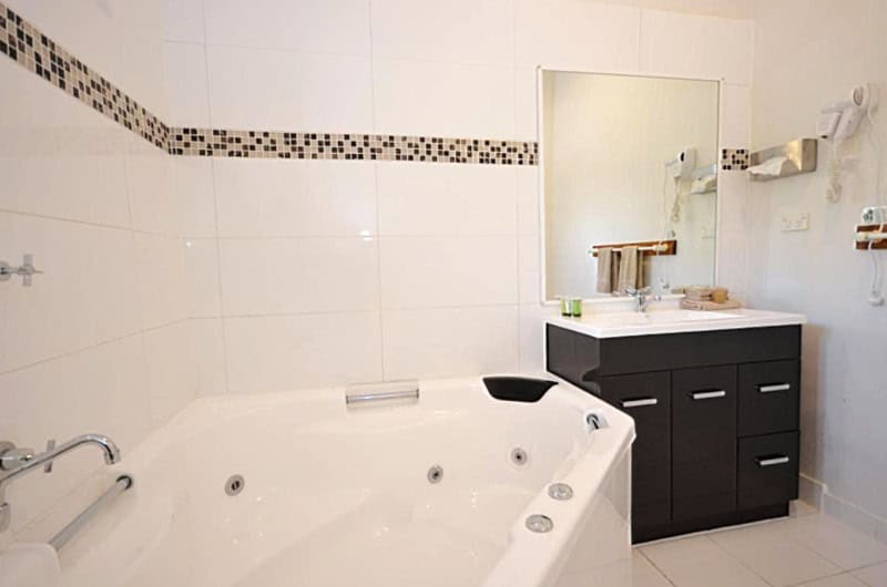 Modern bathroom at Central Springs Inn featuring a large corner spa bath, white tiles with a decorative black and white mosaic tile stripe, and a dark wood vanity with a large mirror above it. The room is equipped with multiple bath amenities including towels and toiletries.