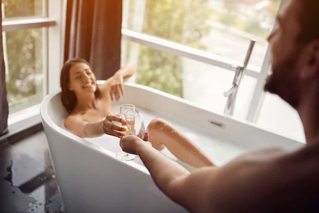 A joyful couple shares a toast with champagne glasses in a luxurious spa bathtub, located in a bright Daylesford accommodation with scenic views from a large window. The man is partially visible in the foreground, while the woman in the tub smiles broadly, enhancing the warm and relaxing atmosphere.