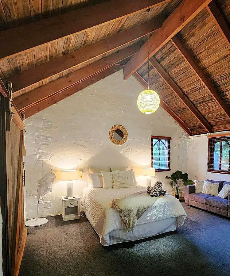 Cozy and inviting bedroom at Daylesford Frog Hollow Estate - The Retreat, featuring a large bed with plush white linens and a beige throw. The room has a high vaulted ceiling with exposed wooden beams, white textured walls, and soft lighting from table lamps and a unique spherical hanging light. A small seating area and decorative plants enhance the tranquil ambiance.