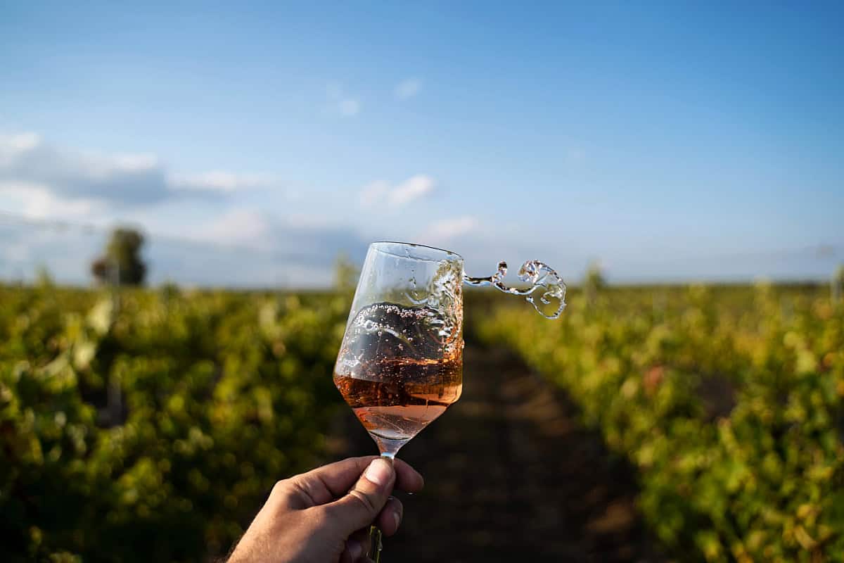 Close-up view of a glass of rosé wine being swirled, with wine splashing out, at a Daylesford wineries vineyard. The background shows rows of lush grapevines under a clear blue sky. The perspective focuses on the dynamic movement of the wine, capturing a moment of enjoyment in a picturesque vineyard setting.