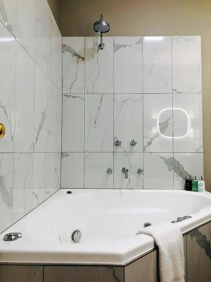 Spacious, modern bathroom at Dudley Boutique Hotel featuring a large white corner spa bath, surrounded by glossy marble tiles with grey veining. An LED mirror and a wall-mounted shower fixture enhance the luxurious feel of the space.