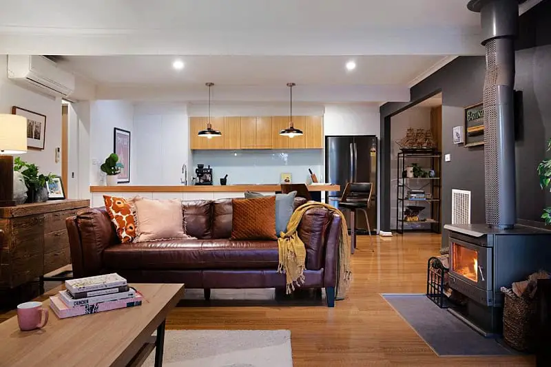 Interior view of the East St Spa House, featuring a cozy living area with a brown leather sofa, decorative throw pillows, and a wooden coffee table. The open-plan layout includes a kitchen with modern appliances and wooden cabinetry, illuminated by pendant lights. A lit fireplace adds warmth to the space.