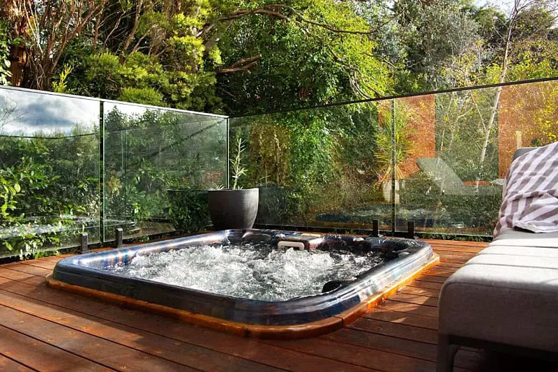 Outdoor spa area at East St Spa House with a bubbling hot tub on a wooden deck, enclosed by clear glass panels. Lush greenery surrounds the area, enhancing the relaxing and private atmosphere.