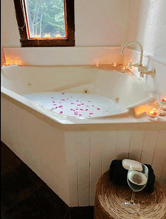 A luxurious spa bath at Frog Hollow Estate - The Retreat, filled with bubbly water and scattered with pink rose petals. The bath is surrounded by flickering candles and a glass of white wine rests on a woven stool, creating a serene and indulgent atmosphere.