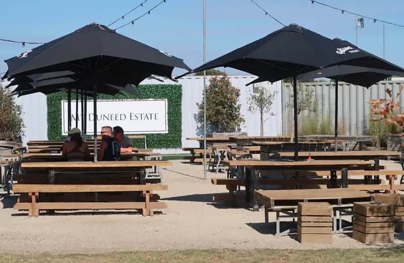 People sitting at wooden picnic tables under large black umbrellas at Mt Duneed Estate winery, with the estate's sign visible in the background. The outdoor seating area is surrounded by a mix of greenery and industrial decor.