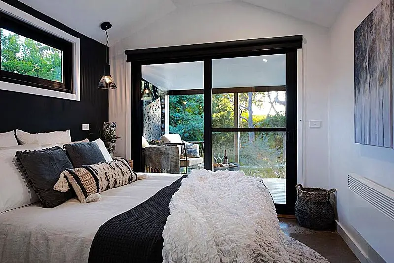 Elegantly designed bedroom at Nest Daylesford accommodation, featuring a large comfortable bed with decorative pillows and a plush throw. Sliding glass doors open to a balcony that looks out onto lush greenery, enhancing the peaceful ambiance. The room is styled with a chic black and white color scheme and modern furnishings.