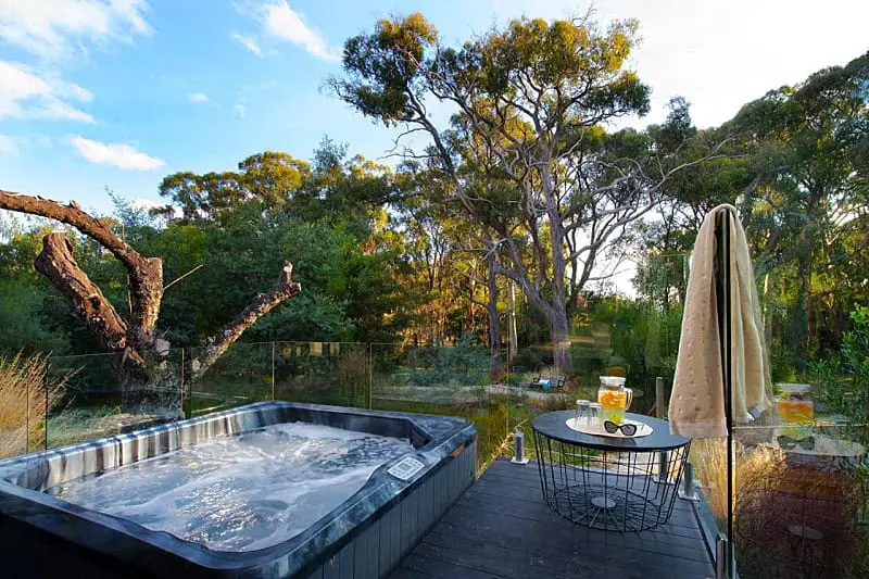 Outdoor area overlooking a serene landscape at Nest Daylesford accommodation with a spa. A bubbling hot tub on a wooden deck with a glass fence features a nearby table hosting a pitcher of lemon water and a towel hanging casually over the chair, all set against a backdrop of lush green trees and clear blue skies.