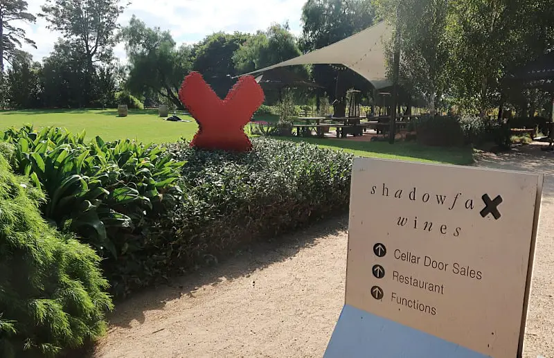 A welcoming scene at Shadowfax Wines features a sign directing visitors to the cellar door sales, restaurant, and functions area. The setting includes a large red "X" sculpture in a landscaped garden, shaded seating areas, and lush greenery under a sunny sky.