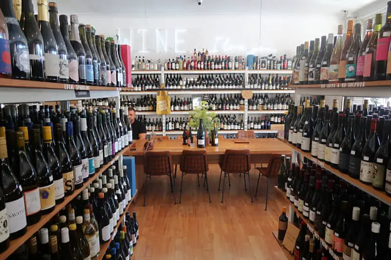 A wide-angle view of Winespeake Cellar + Deli, showcasing rows of wine bottles neatly arranged on wooden shelves with a wooden table and chairs set up for tasting in the center. The walls are lined with an extensive selection of wines, creating a warm and inviting atmosphere.